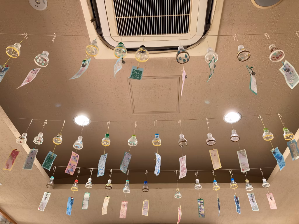 老人ホームの天井に飾られた、入居者や職員によって作られた色鮮やかな短冊が揺れる風鈴の展示。猛暑の中、涼やかな音色を想像させる。