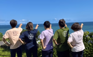 晴れ渡る青空と海をバックに、お揃いのロゴ入りTシャツを着た5人が後ろ姿で一斉に水平線を指さしている様子