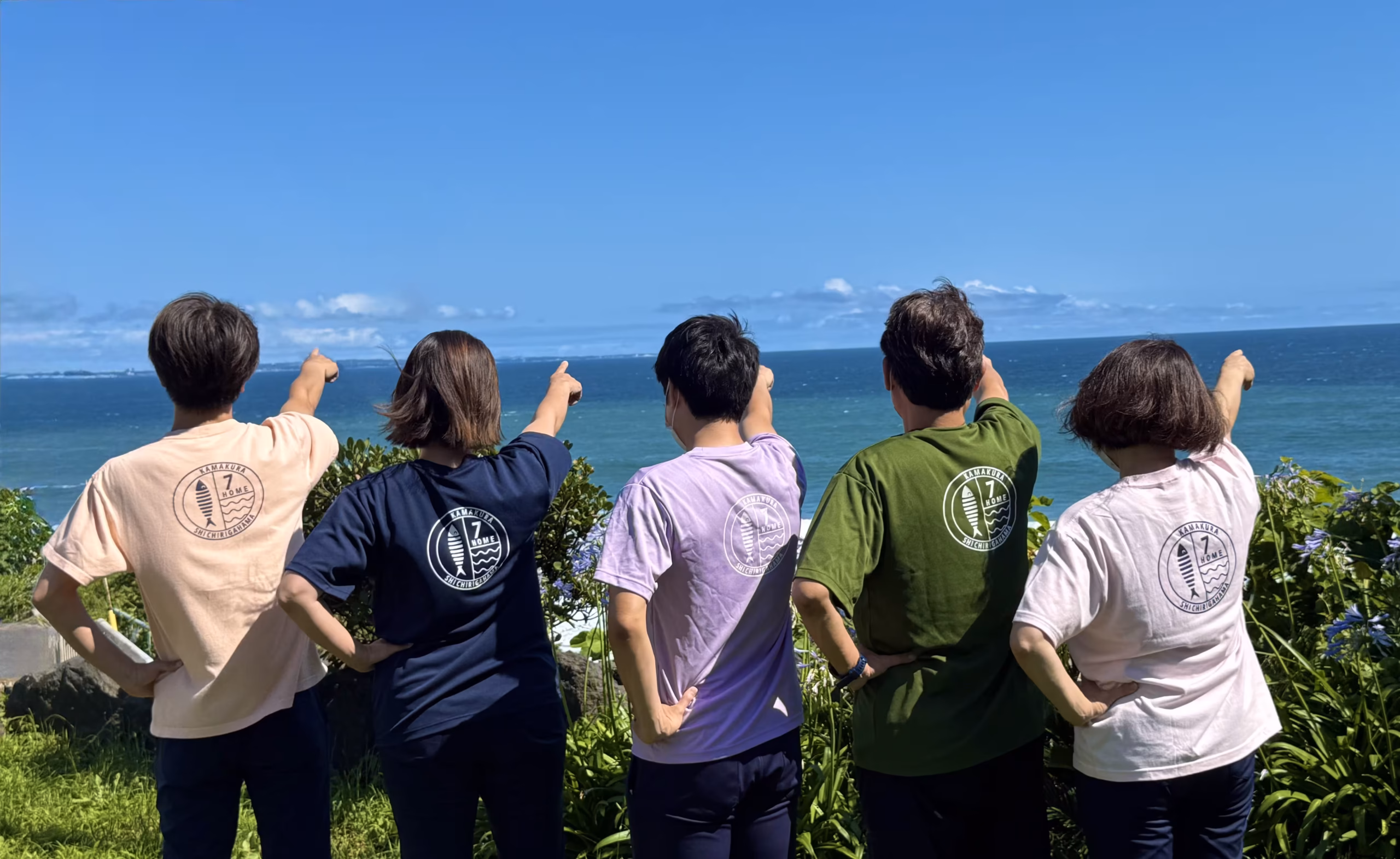 晴れ渡る青空と海をバックに、お揃いのロゴ入りTシャツを着た5人が後ろ姿で一斉に水平線を指さしている様子